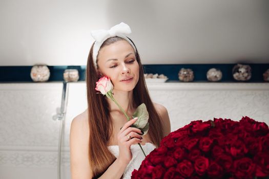 Brunette girl with long hair sitting on bath after morning shower. Pretty lady in white towel looking up and holding flower. Young woman getting bouquet of roses and thinking about boyfriend.