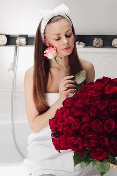 Portrait of gorgeous sensual young brunette woman with headband wrapped in towel sitting in bathroom with bunch of beautiful red roses. She is holding fragile pink rose and smiling at camera. Beauty and skincare concept.