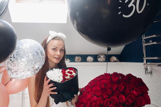 Young girl laughing and enjoying beautiful presents at home. Happy woman sitting neat big balloons and big bouque of red roses, keeping heart of flowers and posing. Concept of happiness and love.