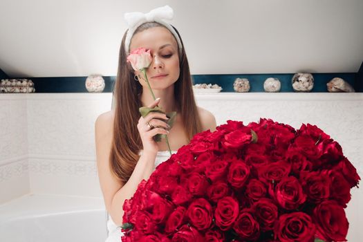 Portrait of gorgeous sensual young brunette woman with headband wrapped in towel sitting in bathroom with bunch of beautiful red roses. She is holding fragile pink rose and smiling at camera. Beauty and skincare concept.