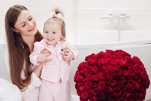 Happy mother and cute daughter sitting in bathroom in morning and smiling. Brunette woman with long hair holding lovely child and looking up. Young wife getting red roses from husband on holiday.