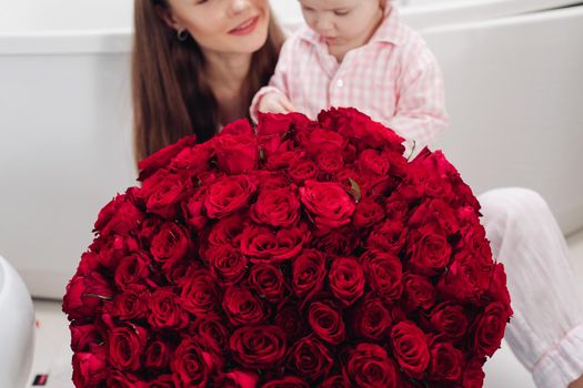 Beautiful young mother posing with little girl and big bouquet of red roses. Pretty woman kissing cute child and smiling. Happy family enjoying present. Concept of love and happiness.