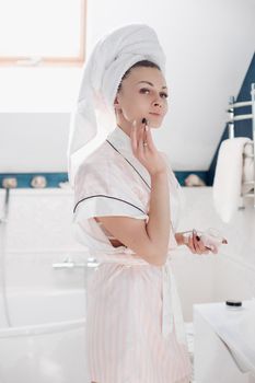 Portrait of cheerful lady in robe and towel on head applying face cream looking at herself in the mirror in the bathroom in the morning. She is smiling while putting cream or lotion on face.