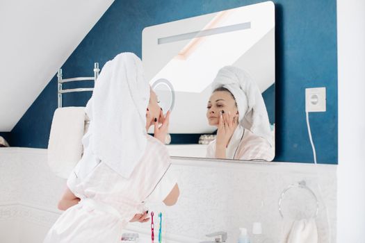 Beautiful woman in white coat and towel on head looking at mirror on her face. Young pretty lady standing in light bathroom after morning shower. Attractive girl using cream on skin before make up.