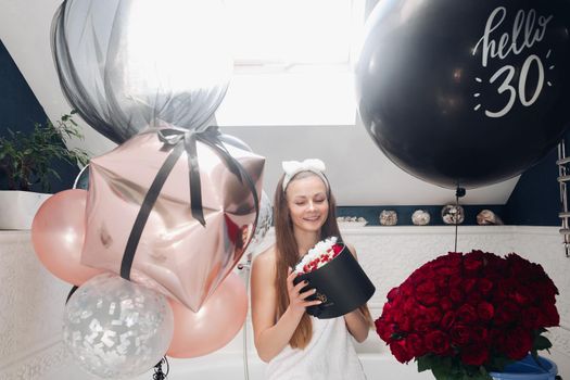 Portrait of positive beautiful brunette woman in headband and towel enjoying flowers in box surrounded by colorful air balloons and bouquet of red roses. She is in the bathroom recieving presents.