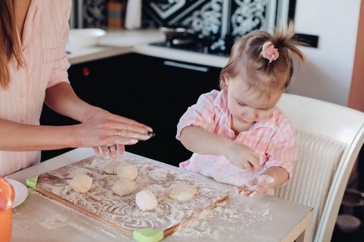 Close up of little serious kid sitting on white chair in pink pajama. Lovely daughter helping her mother preparing breakfast for family. Cute child in flour cooking together with parent at kitchen.