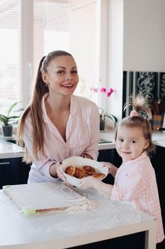 Beautiful woman with lovely child in pajamas holding big bowl with cookies. Close up of happy mother and daughter sitting together at kitchen in morning. Little kid helping parent cooking breakfast.