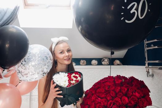 Portrait of positive beautiful brunette woman in headband and towel enjoying flowers in box surrounded by colorful air balloons and bouquet of red roses. She is in the bathroom recieving presents.