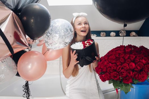 Young girl laughing and enjoying beautiful presents at home. Happy woman sitting neat big balloons and big bouque of red roses, keeping heart of flowers and posing. Concept of happiness and love.