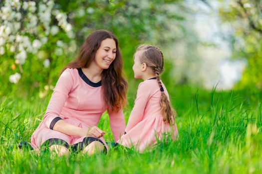 mother and daughter sit in the green grass against the backdrop of blooming apple trees