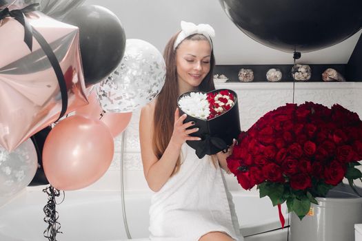 Portrait of positive beautiful brunette woman in headband and towel enjoying flowers in box surrounded by colorful air balloons and bouquet of red roses. She is in the bathroom recieving presents.