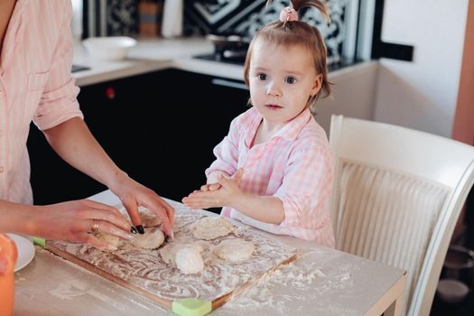 Close up of little serious kid sitting on white chair in pink pajama. Lovely daughter helping her mother preparing breakfast for family. Cute child in flour cooking together with parent at kitchen.
