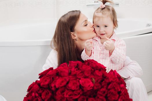 Beautiful young mother posing with little girl and big bouquet of red roses. Pretty woman kissing cute child and smiling. Happy family enjoying present. Concept of love and happiness.