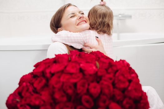 Beautiful young mother posing with little girl and big bouquet of red roses. Pretty woman kissing cute child and smiling. Happy family enjoying present. Concept of love and happiness.