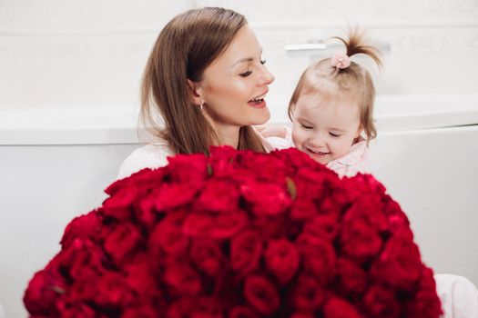 Beautiful young mother posing with little girl and big bouquet of red roses. Pretty woman kissing cute child and smiling. Happy family enjoying present. Concept of love and happiness.