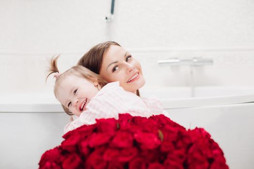 Happy mother and cute daughter sitting in bathroom in morning and smiling. Brunette woman with long hair holding lovely child and looking up. Young wife getting red roses from husband on holiday.