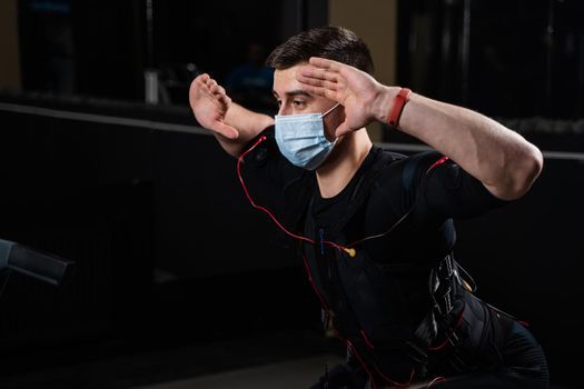 Man in EMS suit and medical mask in gym. Protection from coronavirus covid-19. Sport training in electrical muscle stimulation suit at quarantine period