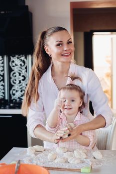 Portrait of gorgeous young adult mother in shirt cooking with her little daughter in similar pink plaid shirt. They are smiling while kneading dough in the kitchen.