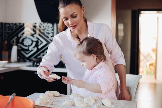 View from side of cheerful woman and cute daughter cooking and baking curd fritter together at home. Lovely family spending time together, laughing and talking in kitchen. Concept of childhood.