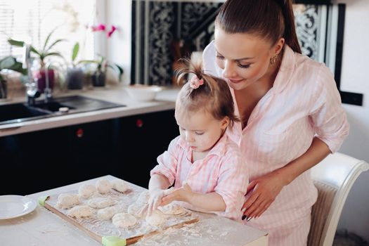 View from side of cheerful woman and cute daughter cooking and baking curd fritter together at home. Lovely family spending time together, laughing and talking in kitchen. Concept of childhood.