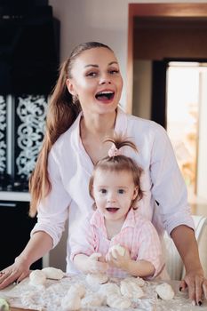 Portrait of gorgeous young adult mother in shirt cooking with her little daughter in similar pink plaid shirt. They are smiling while kneading dough in the kitchen.