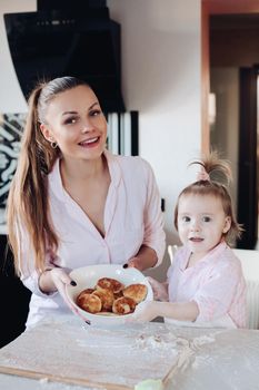 Beautiful woman with lovely child in pajamas holding big bowl with cookies. Close up of happy mother and daughter sitting together at kitchen in morning. Little kid helping parent cooking breakfast.