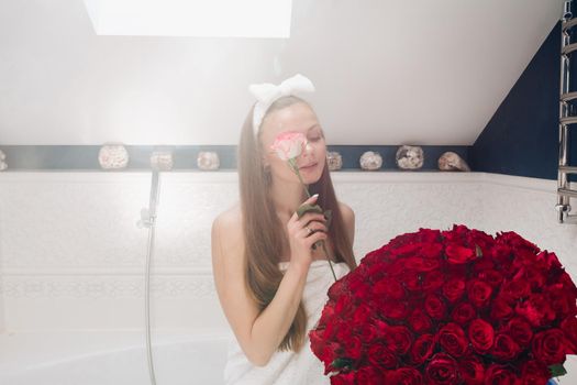 Portrait of gorgeous sensual young brunette woman with headband wrapped in towel sitting in bathroom with bunch of beautiful red roses. She is holding fragile pink rose and smiling at camera. Beauty and skincare concept.