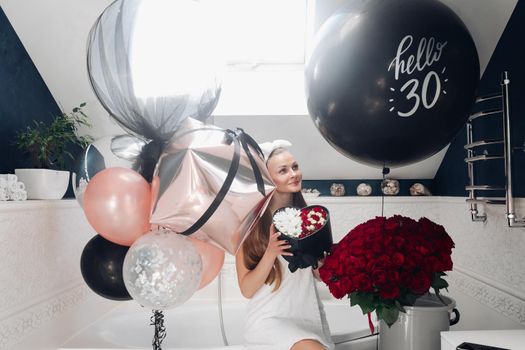 Portrait of positive beautiful brunette woman in headband and towel enjoying flowers in box surrounded by colorful air balloons and bouquet of red roses. She is in the bathroom recieving presents.