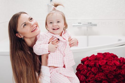 Happy mother and cute daughter sitting in bathroom in morning and smiling. Brunette woman with long hair holding lovely child and looking up. Young wife getting red roses from husband on holiday.