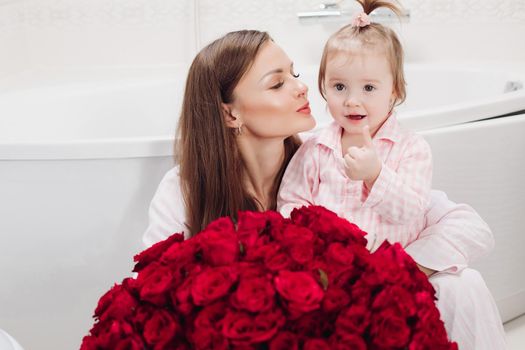 Beautiful young mother posing with little girl and big bouquet of red roses. Pretty woman kissing cute child and smiling. Happy family enjoying present. Concept of love and happiness.