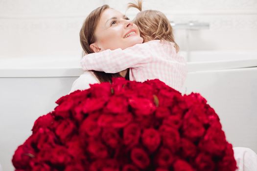 Happy mother and cute daughter sitting in bathroom in morning and smiling. Brunette woman with long hair holding lovely child and looking up. Young wife getting red roses from husband on holiday.