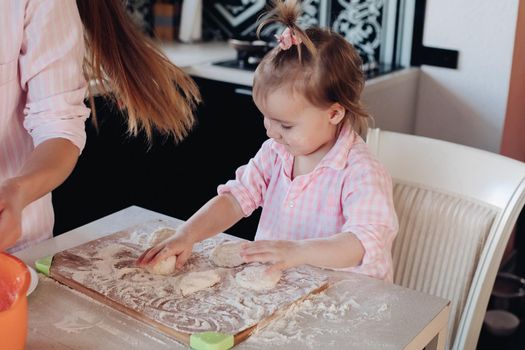 Portrait of lovable child with tail in pink checkered shirt helping mother cooking dumplings or piers in the kitchen. Sitting on chair and kneading dough on board with flour.