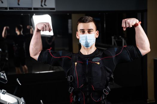 Man in EMS suit and medical mask in gym. Protection from coronavirus covid-19. Sport training in electrical muscle stimulation suit at quarantine period