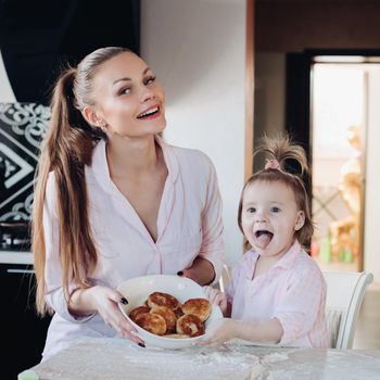 Front view of attractive mother and little daughter looking at camera and showing baked curd fritter. Happy family cooking and playing together in kitchen. Concept of love and happiness.