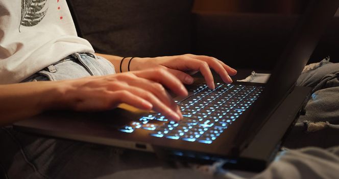 Close up woman hands working typing on keyboard laptop computer at dark room sitting on couch. Freelancer using laptop working from home brightening keyboard.