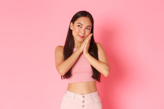 Lifestyle, beauty and women concept. Portrait of beautiful asian girl getting to sleep, lying head on palms but looking away with thoughtful, cunning smile, have interesting plan, pink background.