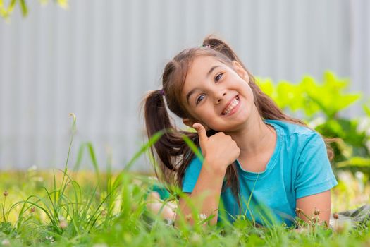 smiling brunette girl with two tails lies on the green grass, on her stomach, in the summer garden, holding thumb up. Copy space