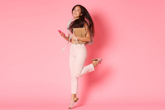 Back to school, education and lifestyle concept. Full length of dreamy and happy, pretty asian girl, female student of university, holding notebooks and smartphone, jumping carefree, pink wall.