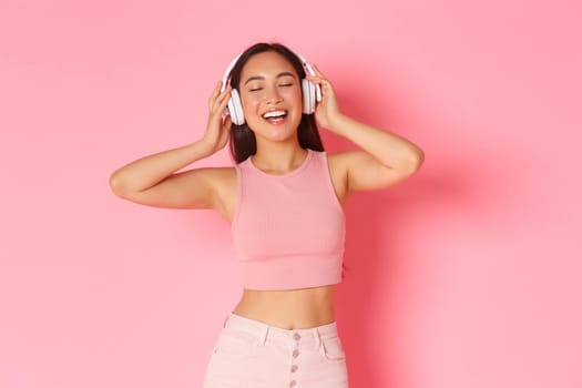Carefree beautiful asian girl in headphones, dancing with eyes closed and relaxed, happy smile, enjoying listening music, hear favorite song, standing over pink background.