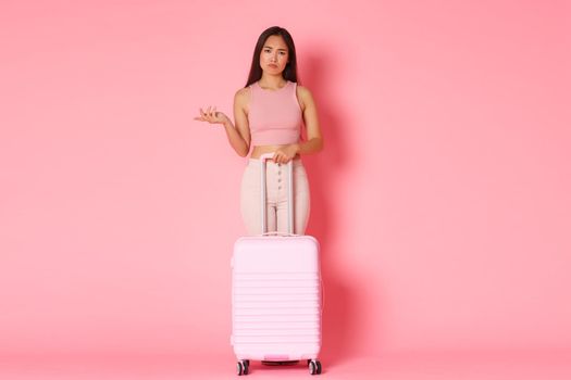 Travelling, holidays and vacation concept. Full-length of frustrated and upset stylish asian girl in summer clothes, raising hand confused, flight canceled over covid-19, stand with suitcase.