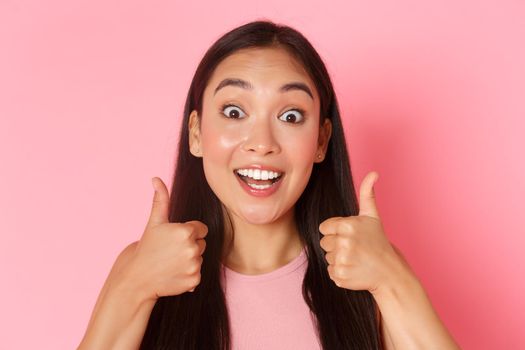 Beauty, fashion and lifestyle concept. Close-up of enthusiastic good-looking asian girl like idea, praising great choice, looking amazed and surprised showing thumbs-up in approval, pink wall.