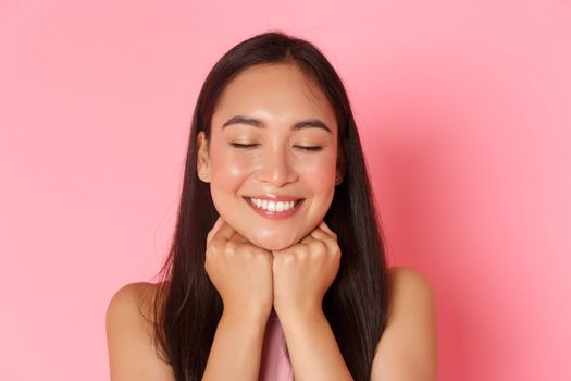 Beauty, fashion and lifestyle concept. Close-up of dreamy and romantic, attractive asian girl daydreaming, leaning face on hands and close eyes with happy white smile, pink background.