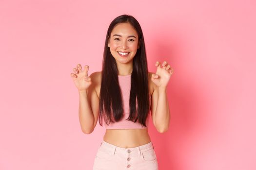 Portrait of silly and cute, upbeat asian girl in trendy summer clothes having fun, acting like lion, making roar gesture with hands mimicking paws, standing over pink background smiling.