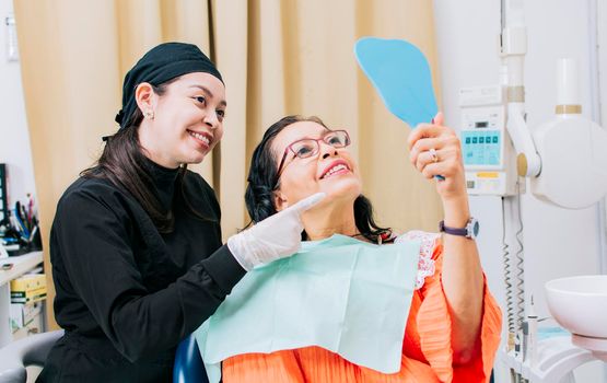 Satisfied female client in dental clinic looking at mirror, Dentist with patient smiling at hand mirror in office, female patient checking teeth after curing teeth in dental clinic,