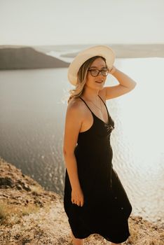 A young woman dress with a hat and glasses stands on a cliff over sea at sunset.