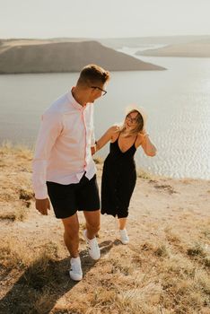 A man and a woman in love walk on a mountain above a large lake by the sea at sunset. A couple of fair-haired fair-skinned people in love are resting in nature in a field