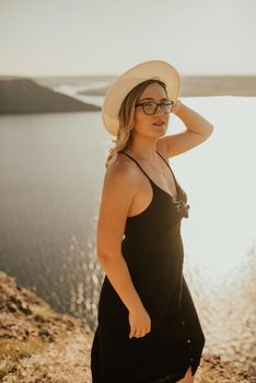 A young woman dress with a hat and glasses stands on a cliff over sea at sunset.