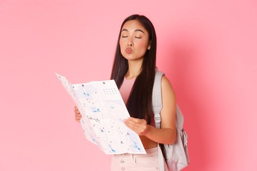 Travelling, lifestyle and tourism concept. Portrait of silly asian girl tourist with backpack and map, close eyes and pouting making kissing face, searching route on vacation, pink background.