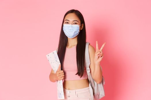 Safe tourism, travelling during coronavirus pandemic and preventing virus concept. Happy asian girl travel in pandemic covid-19 with medical mask, holding map, show peace sign, tourist going abroad.