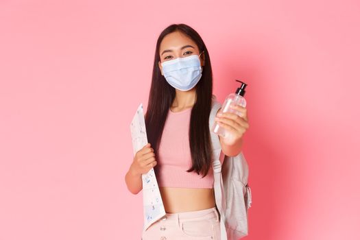 Safe tourism, travelling during coronavirus pandemic and preventing virus concept. Cute asian girl tourist on vacation with medical mask and hand sanitizer, holding map of city, pink background.